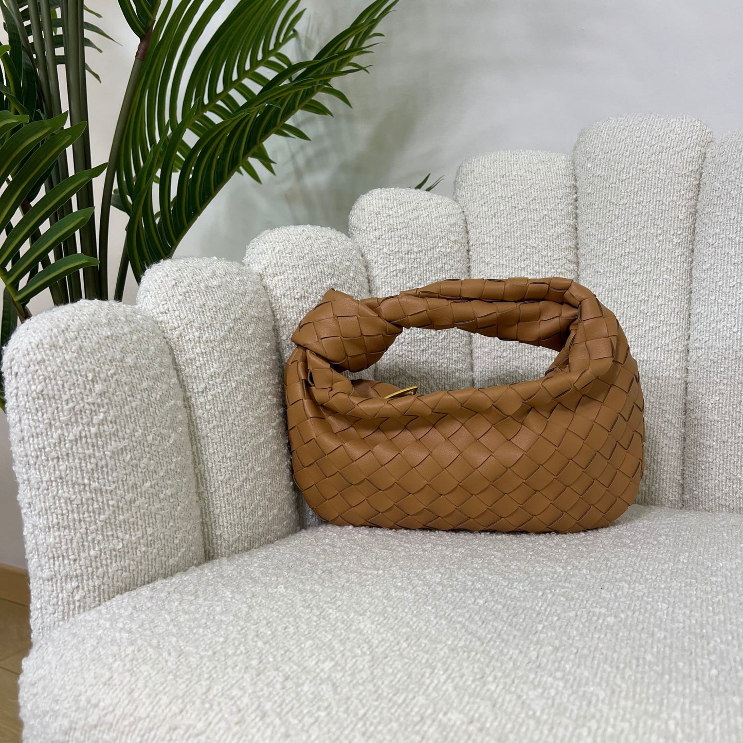 Borsa color cammello intrecciata con manico a nodo, modello Knot ÈCLAT, fotografata su poltrona elegante.